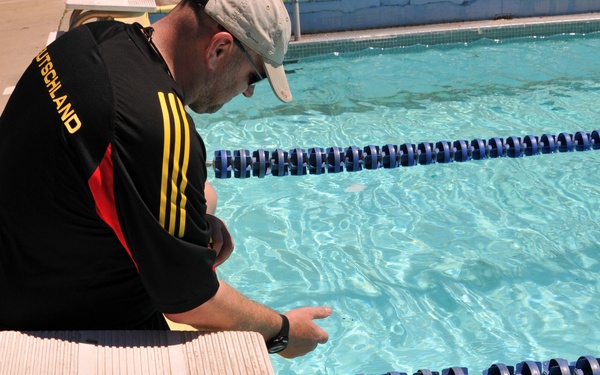 A man wearing a black 'Deutschland' Adidas shirt and a cap crouches at the edge of an outdoor swimming pool, appearing to instruct or assist a person in the water. The swimmer, wearing what appears to be military camouflage clothing, is in the pool and reaching toward the pool edge. The setting is an outdoor lap pool with blue lane dividers.
