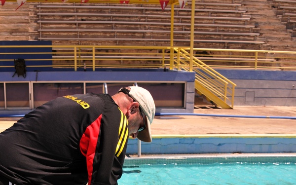 A man in a black athletic shirt and shorts is leaning over the edge of a swimming pool, extending his hand to assist a person in military camouflage uniform who is in the water. The person in the pool appears to be a female soldier being helped out of or supported at the pool's edge. The setting is an outdoor competitive swimming facility with bleachers and lane dividers visible.