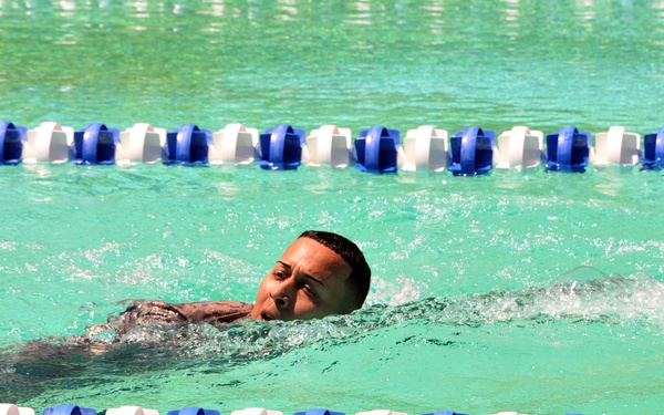 A swimmer is captured mid-stroke in a lap pool, swimming between two lane dividers made of alternating blue and white floats. The water is a clear turquoise-green color, and the swimmer appears to be performing a freestyle or front crawl stroke with their head turned to the side for breathing. The image is taken from a low, water-level angle, emphasizing the swimmer's motion and the surrounding pool environment.