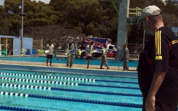 An outdoor swimming pool facility with multiple lap lanes divided by blue lane markers. A man in a black Adidas shirt and cap stands poolside observing a swimmer in the water. In the background, several individuals in military-style uniforms are visible near vehicles and pool deck areas.