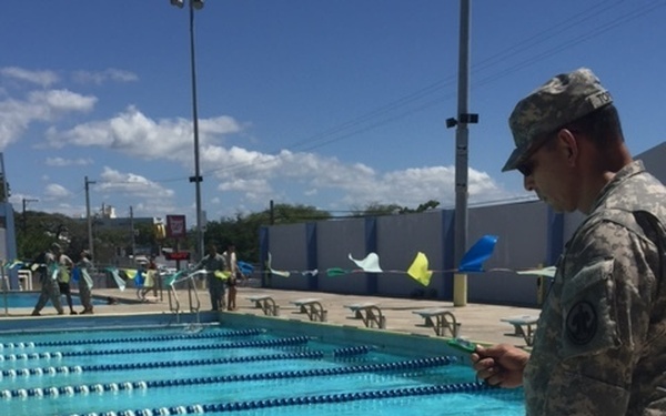 A military personnel in Army Combat Uniform (ACU) pattern camouflage stands at the edge of an outdoor swimming pool, appearing to time or monitor swimmers in the water. At least two swimmers in military uniforms are visible in separate lanes of the pool. The setting appears to be a military swim qualification or fitness assessment event at an outdoor aquatic facility.