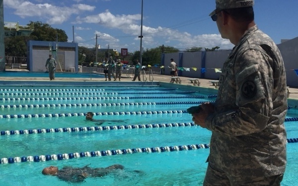 A military swim assessment or training exercise is taking place at an outdoor pool facility. Soldiers in full combat uniforms (ACU pattern) are swimming in the pool lanes while a uniformed observer holding a clipboard monitors and records their performance from the pool deck. Additional military personnel are visible in the background near the pool area.
