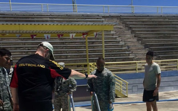 A group of individuals, including several in military camouflage uniforms, are gathered at an outdoor swimming pool. One person wearing a black 'Deutschland' jersey appears to be instructing or supervising, while a swimmer in camouflage clothing performs a backstroke in the pool. The setting appears to be a military swim training or assessment event at a facility with stadium-style bleachers.