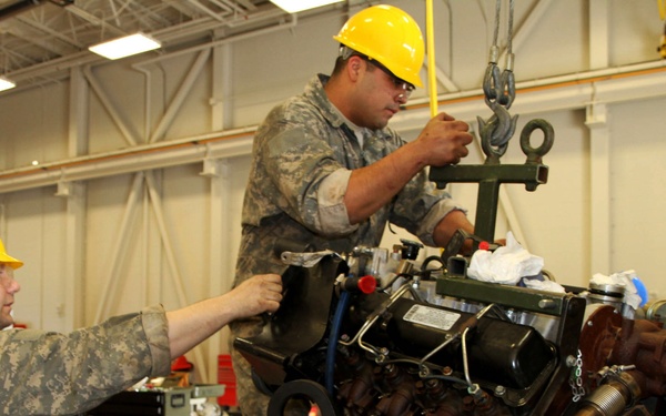 Military personnel in camouflage uniforms and yellow hard hats are working in a maintenance bay, performing an engine removal or installation using an overhead crane/hoist system. At least three individuals are visible working collaboratively on what appears to be a large vehicle engine. The setting is an indoor military motor pool or maintenance facility equipped with standard workshop tools and equipment.
