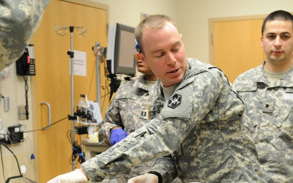 Military medical personnel in U.S. Army uniforms are performing or training on emergency medical procedures on a patient lying on a stretcher. The setting appears to be a military medical facility or trauma bay, with multiple IV lines, blood bags, and medical equipment visible. This may be a training exercise or an actual medical emergency response scenario.