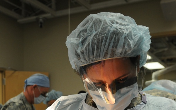 A surgical team is performing an operation in what appears to be a military medical facility. The primary surgeon in the foreground is wearing full sterile surgical attire including a blue cap, face shield, surgical mask, and yellow latex gloves, and is actively working with surgical instruments. A second individual in military camouflage uniform and surgical cap is visible in the background.