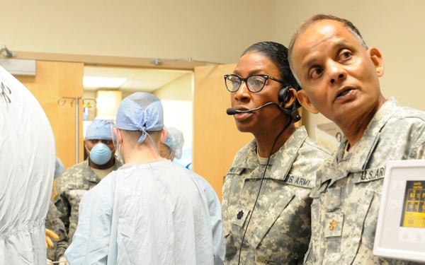 The image shows a military medical setting where U.S. Army personnel in ACU (Army Combat Uniform) are observing or overseeing a medical procedure. Two soldiers in uniform stand near medical equipment while medical staff in surgical gowns and masks perform or assist with a procedure in the background. The setting appears to be a hospital operating or procedure room.