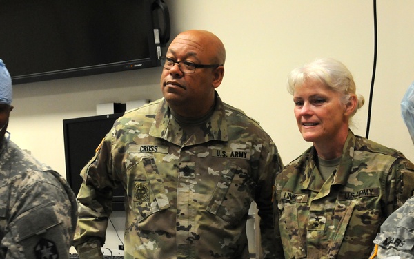 Four U.S. Army personnel are gathered in what appears to be a medical training or clinical facility. Two individuals are wearing surgical masks and gloves, suggesting active medical procedures or training, while two others in standard Army Combat Uniforms (ACU/OCP) appear to be observing. Name tapes visible include 'CROSS' and 'LINK' on OCP uniforms, and a partially visible name on the left.