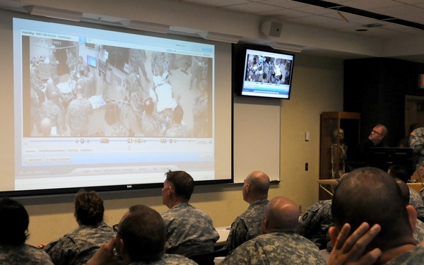 Military personnel in Army Combat Uniform (ACU) digital camouflage pattern are seated in a classroom-style briefing room, attending a presentation. A large projection screen displays what appears to be aerial or overhead imagery, possibly of a combat or disaster zone, alongside a software interface. Additional flat-screen monitors are mounted on the walls showing similar content.