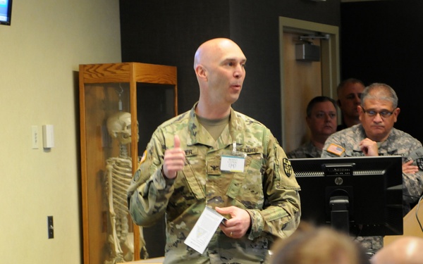 A bald U.S. Army soldier in MultiCam/OCP camouflage uniform is presenting or briefing to a seated audience of military personnel in a classroom or training facility. An anatomical skeleton model is visible in a display case behind the speaker, suggesting a medical or health-related training environment. Multiple uniformed personnel from what appear to be both Army and Air Force branches are seated as attendees.