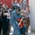 A person in blue digital camouflage military uniform and tactical face mask is aiming a rifle toward a door or hatch on the right side of the frame. They are surrounded by a large group of media personnel — photographers and videographers — many wearing orange press vests, actively documenting the scene. This appears to be a staged military demonstration or exercise being covered by press.