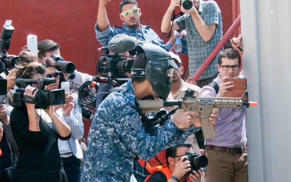 A person in blue digital camouflage military uniform and tactical face mask is aiming a rifle toward a door or hatch on the right side of the frame. They are surrounded by a large group of media personnel — photographers and videographers — many wearing orange press vests, actively documenting the scene. This appears to be a staged military demonstration or exercise being covered by press.