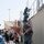 The image depicts a military training or demonstration exercise, likely a boarding or vessel seizure drill, being conducted in front of an audience of media and observers. Personnel in U.S. Navy blue digital camouflage uniforms and protective face masks are climbing a metal staircase alongside what appears to be a simulated ship structure made of shipping containers. The exercise is being photographed and filmed by a large crowd of journalists and spectators.