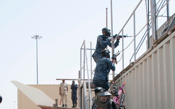 The image depicts a military training or demonstration exercise, likely a boarding or vessel seizure drill, being conducted in front of an audience of media and observers. Personnel in U.S. Navy blue digital camouflage uniforms and protective face masks are climbing a metal staircase alongside what appears to be a simulated ship structure made of shipping containers. The exercise is being photographed and filmed by a large crowd of journalists and spectators.