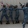 A group of personnel in blue digital camouflage uniforms and protective helmets/masks are conducting a tactical training exercise. They are moving in a stack formation along a wall, each holding training rifles with orange/red muzzle tips indicating non-lethal or airsoft weapons. The exercise appears to be a close-quarters battle (CQB) or room-clearing drill.