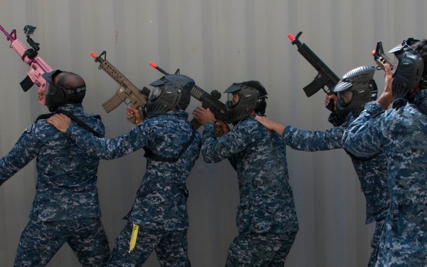 A group of personnel in blue digital camouflage uniforms and protective helmets/masks are conducting a tactical training exercise. They are moving in a stack formation along a wall, each holding training rifles with orange/red muzzle tips indicating non-lethal or airsoft weapons. The exercise appears to be a close-quarters battle (CQB) or room-clearing drill.