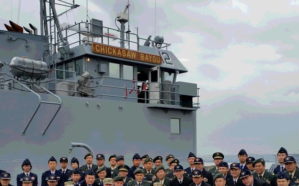 A large group of military personnel and civilians pose for a formal group photograph in front of the US Army vessel 'Chickasaw Bayou' at a dock or port facility. The group includes members wearing various military uniforms from what appear to be multiple nations, including US Army camouflage, dark naval uniforms, and green military dress uniforms. The setting suggests a multinational military visit, exchange, or ceremony at a naval installation.