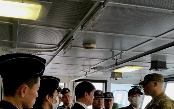 A group of military and naval personnel from what appears to be multiple services are gathered inside a ship's bridge or interior compartment. The group includes individuals in dark naval uniforms, olive/green military uniforms, and one person in US Army multicam/OCP combat uniform, suggesting a multinational military meeting or tour. The setting appears to be aboard a naval vessel, indicated by the ship's windows, overhead piping, and industrial interior.