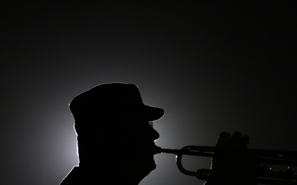 A silhouetted figure wearing a peaked cap is playing a brass instrument, likely a bugle or trumpet, against a dark nighttime background with a single diffused light source behind them. The dramatic backlighting creates a stark silhouette effect, obscuring all facial and clothing details. The composition suggests a ceremonial or military context given the cap style and instrument choice.