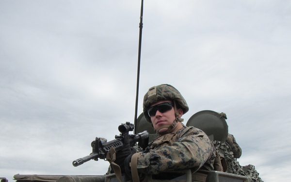 A soldier in digital camouflage uniform is positioned in the hatch of an armored military vehicle, holding a rifle with an optical scope. The soldier is wearing a combat helmet, tactical sunglasses, and gloves, appearing to be in a security or overwatch posture. A radio antenna extends vertically from the vehicle, and overcast skies are visible in the background.