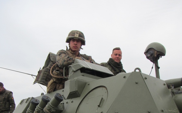 The image shows military personnel on top of a green armored fighting vehicle, likely during a joint training exercise. Two soldiers in what appears to be US Marine Corps camouflage uniforms are positioned in or near the turret hatch, while a third soldier in a different camouflage pattern (possibly Spanish or another NATO ally) is visible on the left side of the vehicle. The setting appears to be an open, hilly terrain under an overcast sky.