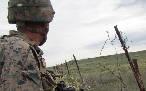 A military service member in MARPAT digital camouflage uniform is positioned near a barbed wire fence line, observing an open grassland field. The soldier is equipped with a rifle featuring an optic sight and is carrying a tactical backpack. The setting appears to be a rural training or operational environment under overcast skies.