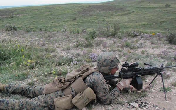 A military service member in MARPAT (Marine pattern) camouflage uniform is in a prone firing position on open terrain. The soldier is aiming what appears to be an M249 Squad Automatic Weapon (SAW) with a bipod deployed on rocky, semi-arid ground. The setting appears to be a training exercise or field operation in a grassland/scrubland environment.
