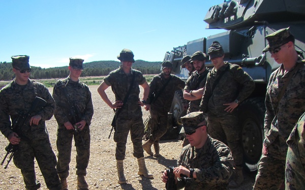 A group of military personnel in MARPAT (Marine digital camouflage) uniforms are gathered outdoors in an arid training environment. One Marine is kneeling on a brown cloth/mat, demonstrating weapon disassembly or maintenance to the surrounding group. A large military vehicle is visible in the background to the right.