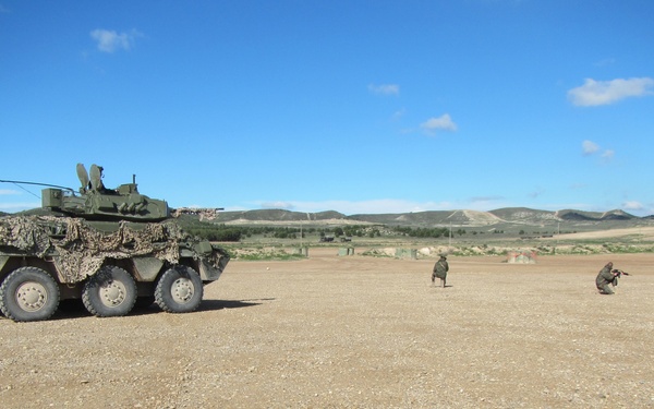 A military training exercise is taking place on an open, arid terrain. A wheeled armored fighting vehicle, covered with camouflage netting, is positioned on the left side, while two soldiers in combat gear are visible in the middle distance performing tactical maneuvers. The setting appears to be a military training ground with rolling hills and sparse vegetation in the background.
