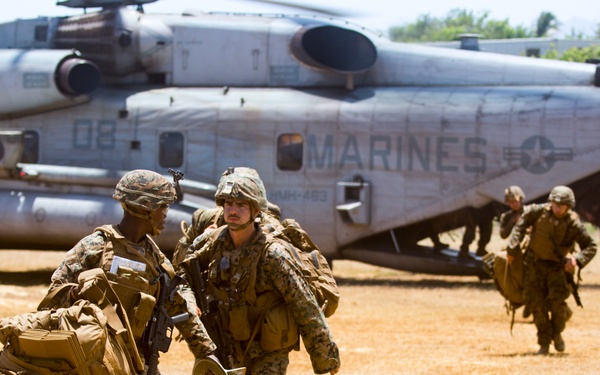 A group of U.S. Marines are disembarking from a large military helicopter, identified as a CH-53E Super Stallion marked 'MARINES' and numbered '08', on a dry, open field. The Marines are in full combat gear, carrying heavy packs and equipment, moving away from the aircraft in an organized manner. The scene appears to depict a training exercise or tactical insertion operation.