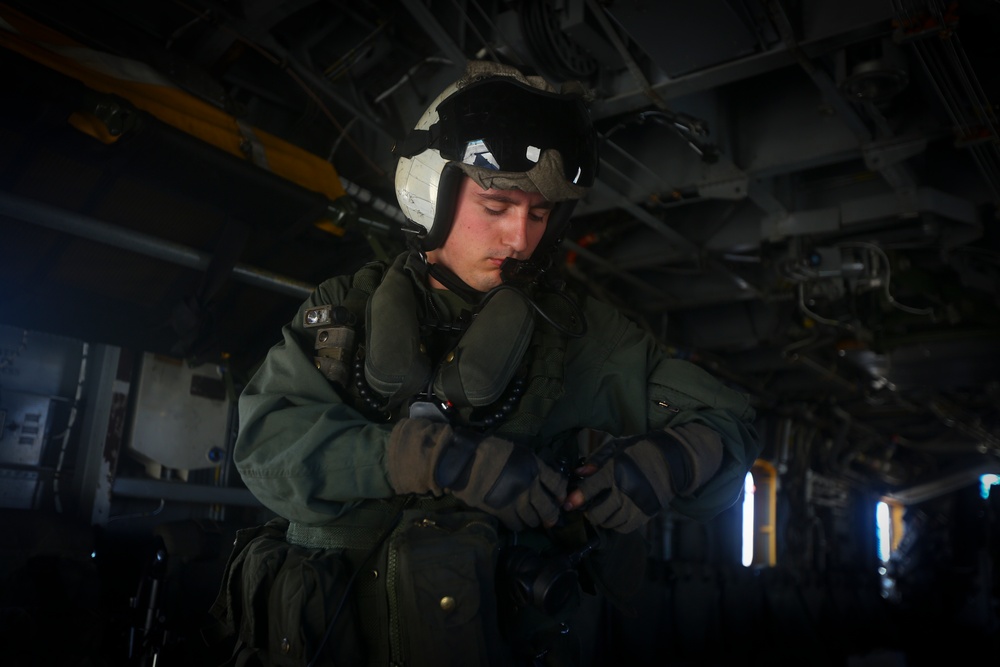 A military aviator or aircrew member is photographed inside what appears to be a military aircraft, likely a helicopter or transport aircraft. The individual is wearing full flight gear including a flight helmet with visor, oxygen mask, flight suit, and tactical gloves. The person appears to be checking or adjusting their equipment while standing in the aircraft's interior.