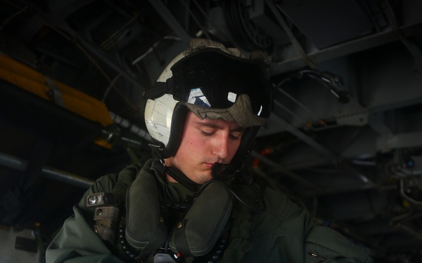 A military aviator or aircrew member is photographed inside what appears to be a military aircraft, likely a helicopter or transport aircraft. The individual is wearing full flight gear including a flight helmet with visor, oxygen mask, flight suit, and tactical gloves. The person appears to be checking or adjusting their equipment while standing in the aircraft's interior.