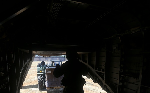 The image is taken from inside a military cargo aircraft looking outward through the open rear ramp. A silhouetted figure stands on the ramp while a forklift approaches carrying a pallet of supplies. The scene appears to depict a cargo loading or humanitarian aid operation at a dirt airstrip.