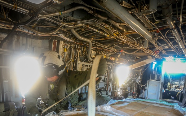 The image shows the interior cargo hold of a military transport aircraft, likely a C-130 Hercules or similar heavy-lift aircraft. A crew member wearing a flight helmet and military flight suit is visible working near secured cargo pallets wrapped in plastic and cardboard. The cargo is strapped down with heavy-duty tie-down straps, and the aircraft's exposed wiring, hydraulic lines, and structural components are visible along the ceiling and walls.