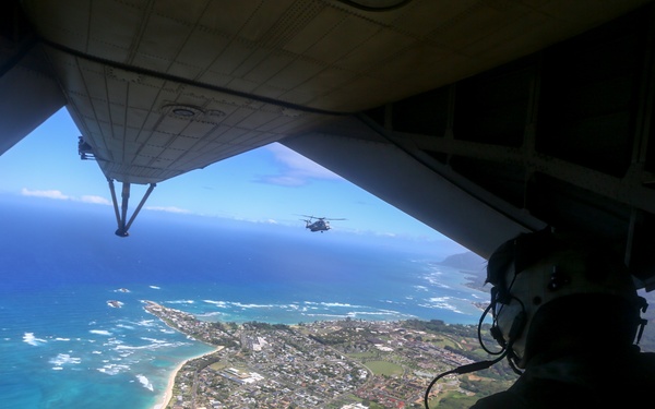 An aerial military photograph taken from the open rear ramp of a large military transport aircraft, showing a crew member in flight gear seated at the edge overlooking a coastal town and turquoise ocean below. A second military helicopter is visible flying in formation in the distance. The landscape below appears to be a Hawaiian island, identifiable by the distinctive coastline, vegetation, and turquoise water.