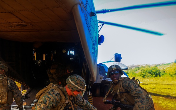 Military personnel in MARPAT (Marine pattern) camouflage uniforms are unloading cardboard boxes from a large helicopter onto a red dirt landing zone. The soldiers are armed and equipped with tactical gear while conducting what appears to be a humanitarian aid or logistics resupply operation. The scene takes place in a rural, vegetated area with red soil terrain.