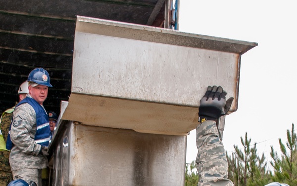 A group of military personnel and civilian workers are collaboratively unloading or moving a large stainless steel industrial sink or equipment from the back of a truck. Personnel are wearing a mix of US Air Force digital camouflage uniforms and civilian work attire, with safety vests and hard hats indicating a structured work operation. The scene appears to be a humanitarian assistance, disaster relief, or joint training exercise conducted in a wooded outdoor environment.