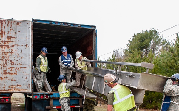 A group of military personnel and workers in safety vests and hard hats are unloading a large stainless steel commercial sink unit from a blue cargo trailer truck. The team appears to be conducting a humanitarian aid, disaster relief, or community support operation, with members wearing a mix of military uniforms (Air Force ABUs, Marine MARPAT) and high-visibility safety gear. The setting is a rural or semi-wooded area with muddy ground conditions.