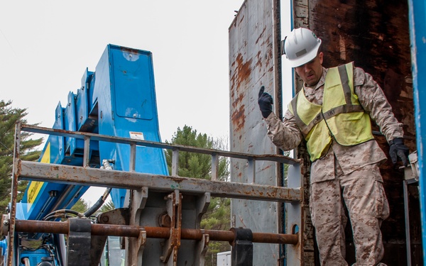 A military service member in camouflage uniform, white hard hat, and high-visibility yellow safety vest is working on or near a flatbed trailer at what appears to be a military camp. A large blue telescoping boom lift or telehandler is positioned nearby. The setting appears to be a wooded military installation, with a sign partially reading 'CAMP' visible in the background.