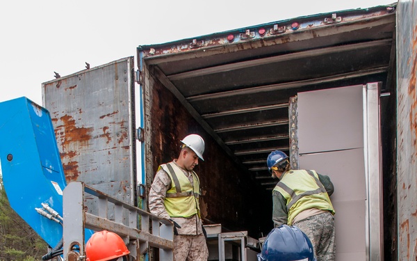 Four military personnel in camouflage uniforms are unloading or loading equipment from a rusted semi-trailer truck at an outdoor location. The personnel are wearing high-visibility safety vests and hard hats in various colors (white, blue, orange/red). Two service members appear to be in Marine Corps MARPAT uniforms while at least one is in Air Force ABU pattern.