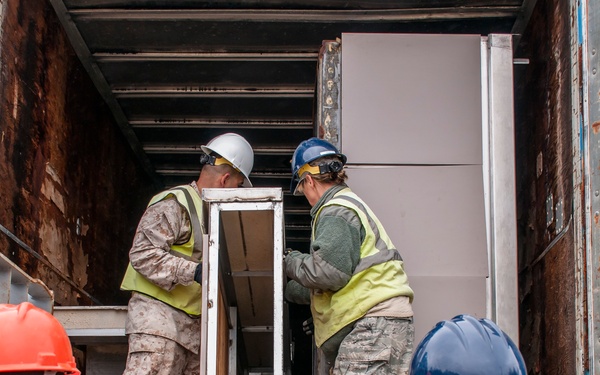 Military personnel in camouflage uniforms and civilian workers wearing high-visibility safety vests and hard hats are collaborating to load or unload equipment into a heavily rusted shipping container or trailer. The team appears to be handling a white-framed piece of equipment or furniture. This appears to be a logistics or humanitarian aid operation involving joint military and civilian coordination.