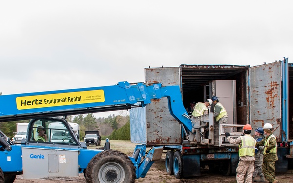 A blue Hertz Equipment Rental telehandler (Genie brand) is being used to unload or load equipment from a shipping container on a flatbed trailer at a muddy outdoor site. Several workers, including individuals in military camouflage uniforms and civilian safety vests with hard hats, are coordinating the operation. The setting appears to be a field or staging area surrounded by pine trees under overcast skies.