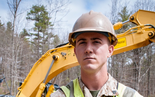 A young male in U.S. Marine Corps desert camouflage uniform stands with arms crossed wearing a tan hard hat and high-visibility safety vest. He is positioned in front of a large yellow Hertz Equipment Rental excavator at what appears to be a construction or engineering site. The setting is a cleared area surrounded by bare deciduous trees, suggesting early spring conditions.