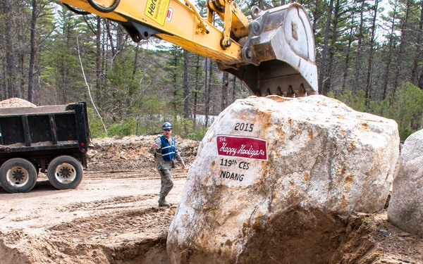A large boulder is being lifted by a yellow excavator at a construction or land-clearing site surrounded by pine trees. The boulder has a painted sign reading '2015 / The Happy Hooligans / 119th CES / NDANG', indicating it is associated with the 119th Civil Engineer Squadron of the North Dakota Air National Guard. A worker in military-style uniform and hard hat stands nearby next to a black dump truck.