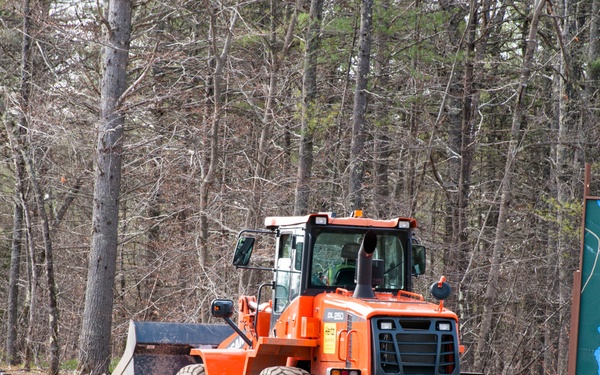 An orange front-end loader/wheel loader is operating on a dirt road at the entrance of Camp William Hinds, a Boy Scouts of America camp located near Panther Pond. Two workers wearing hard hats and high-visibility vests are walking alongside the machinery on the unpaved road. The scene appears to be a construction or grounds maintenance operation during early spring, as indicated by the bare deciduous trees in the background.
