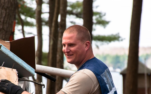 A man wearing a blue safety/reflective vest over a tan t-shirt is handling debris or construction materials in an outdoor wooded setting near a body of water. He is smiling and wearing black work gloves while managing what appears to be a pile of mixed debris including wood, metal, and other materials. The background shows tall pine trees, a lake, and a small structure in the distance.