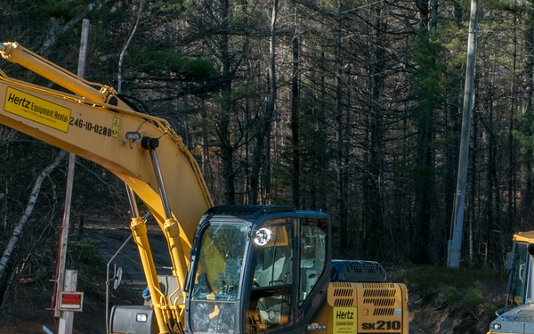 Seven military personnel in camouflage uniforms pose together in front of two pieces of yellow Hertz Equipment Rental construction machinery at what appears to be a training or work site. The group stands between a large tracked excavator (SK210) and a bulldozer on a dirt clearing surrounded by pine trees. The image appears to document a military engineering or construction training exercise.