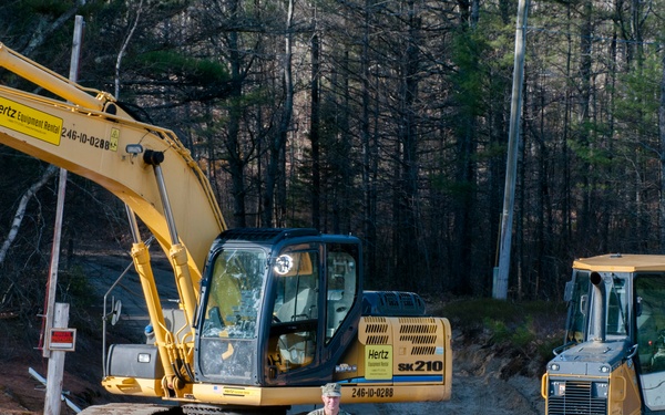 A construction site in a wooded area features two pieces of yellow Hertz Equipment Rental machinery: a tracked excavator (model SK210) and a bulldozer. A person in military-style camouflage uniform stands between the two machines on a dirt surface. A speed limit sign is partially visible on the right side near a utility pole.
