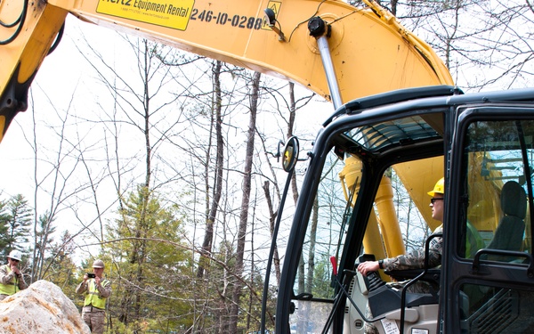 A yellow Hertz Equipment Rental excavator (unit number 246-10-0280) is being operated at an outdoor construction or land-clearing site surrounded by trees. The operator, wearing a hard hat and camouflage clothing, sits in the cab while two other workers in similar attire observe from a distance. Large rocks and disturbed earth are visible in the foreground.