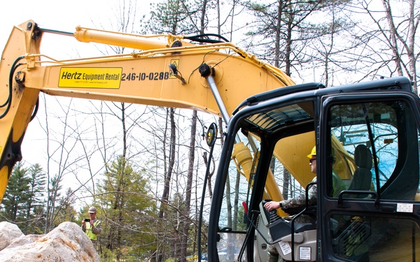 A yellow Hertz Equipment Rental excavator (unit number 246-10-0288) is actively being operated at a construction or land-clearing site surrounded by trees. An operator is seated in the cab while a second worker in a hard hat and safety vest stands nearby observing. Large rocks and disturbed earth are visible in the foreground, indicating active excavation work.